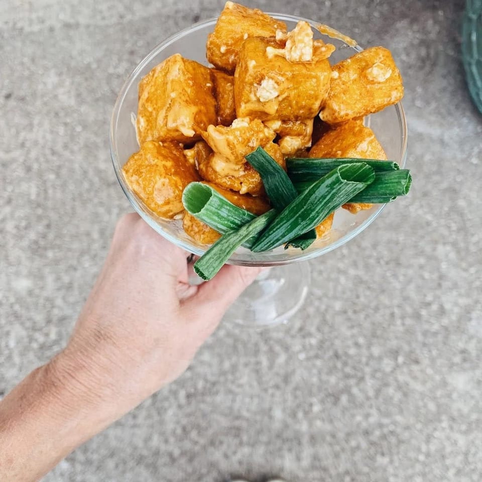 A hand holding a glass filled with golden-brown tofu cubes and green onion garnishes.