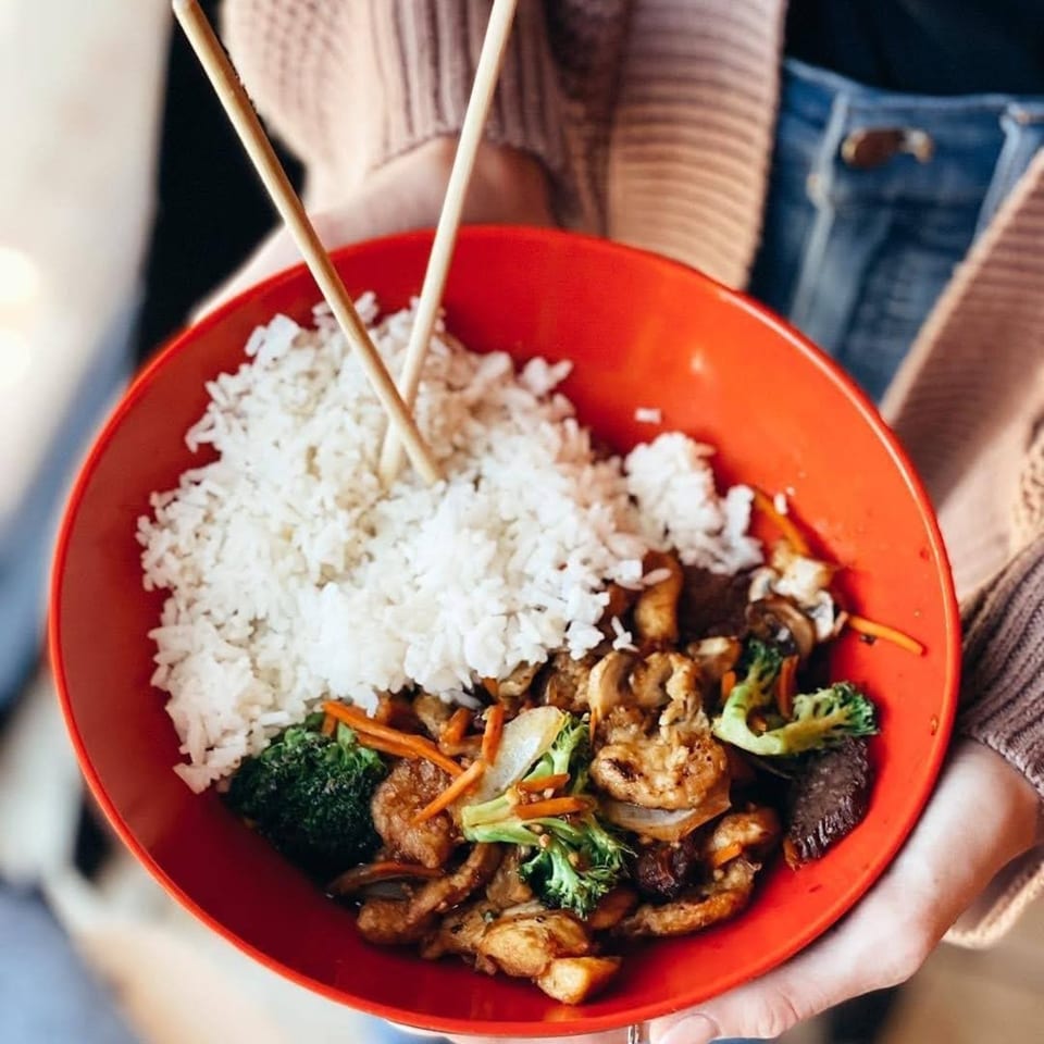 Hands holding a red bowl of Leo's Asian Bistro stir-fry with white rice and chopsticks.