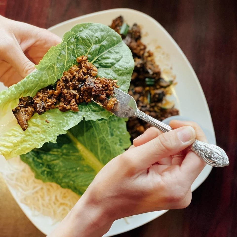 Hands preparing a lettuce wrap with savory Asian-fusion filling and noodles on a white plate.