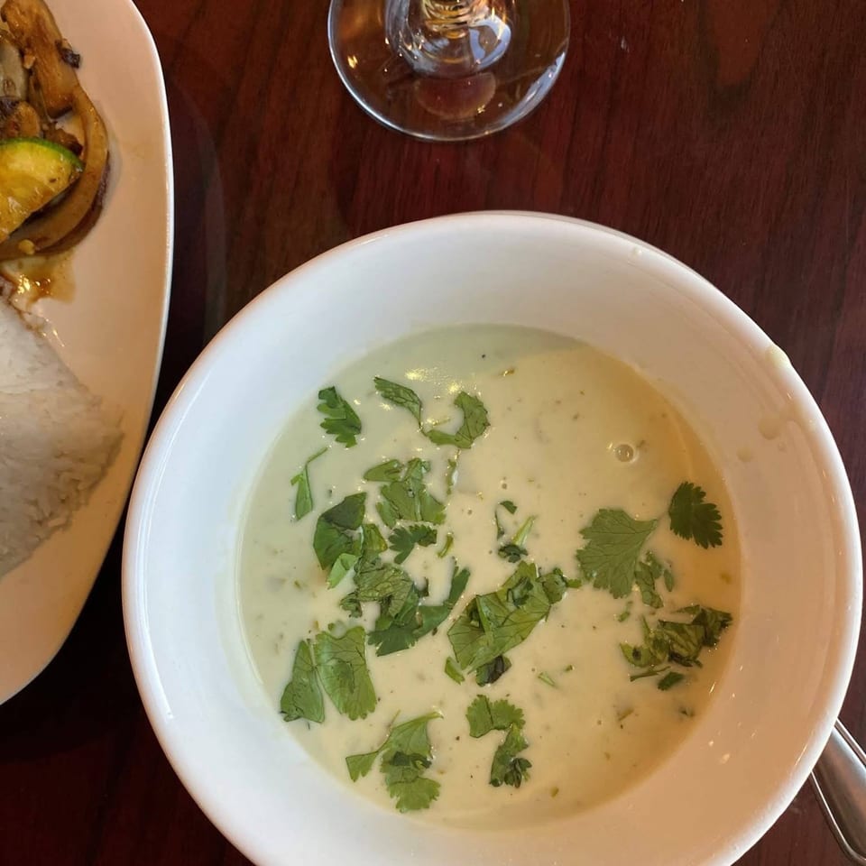 A white bowl of creamy green soup with cilantro, next to a plate of food and a wine glass.